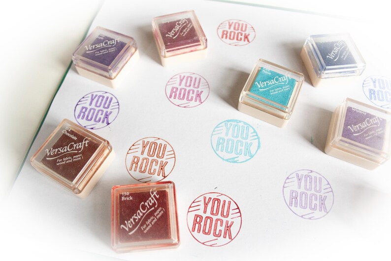 K&ouml;nnte beinhalten: Eine Sammlung von neun Stempelfarben mit dem aufgedruckten Text "You Rock". Die Stempelfarben sind in verschiedenen Farben, darunter Rot, Blau, Gr&uuml;n und Lila. Die Stempelfarben sind in einem Rastermuster auf einer wei&szlig;en Oberfl&auml;che angeordnet.