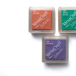 K&ouml;nnte beinhalten: Drei Stempelkissen in durchsichtigen Plastikbeh&auml;ltern. Die Stempelkissen sind mit "VersaCraft" beschriftet und haben die Farben Aprikose, Flieder und Mintgr&uuml;n. Die Stempelkissen sind f&uuml;r Stoff, Papier, Holz und mehr.