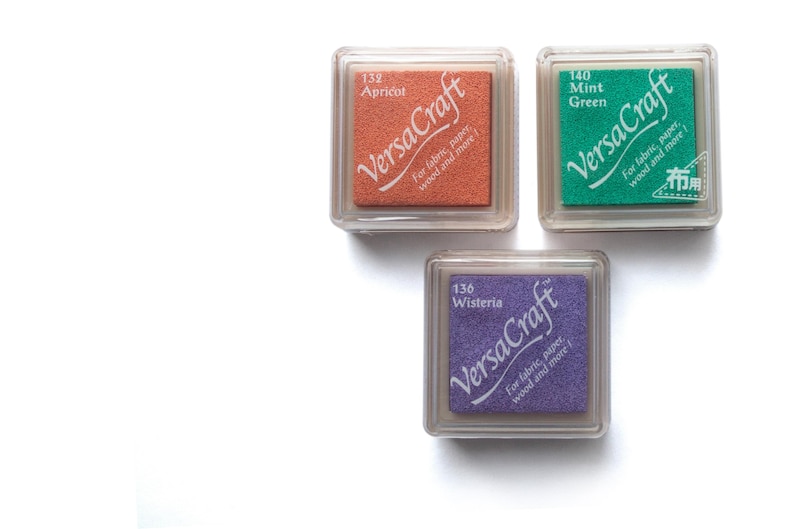 K&ouml;nnte beinhalten: Drei Stempelkissen in durchsichtigen Plastikbeh&auml;ltern. Die Stempelkissen sind mit "VersaCraft" beschriftet und haben die Farben Aprikose, Flieder und Mintgr&uuml;n. Die Stempelkissen sind f&uuml;r Stoff, Papier, Holz und mehr.