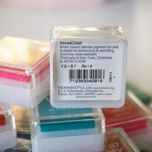 Pode incluir: Uma almofada de tinta VersaColor na cor dourada. A almofada de tinta est&aacute; em um recipiente de pl&aacute;stico transparente com um r&oacute;tulo branco. O r&oacute;tulo inclui o nome da marca, a cor e as informa&ccedil;&otilde;es do produto. A almofada de tinta &eacute; livre de &aacute;cidos, resistente ao desbotamento, segura para crian&ccedil;as e n&atilde;o t&oacute;xica.