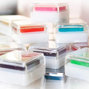 Pode incluir: Uma cole&ccedil;&atilde;o de almofadas de tinta de pl&aacute;stico transparente com almofadas de tinta coloridas dentro. As almofadas de tinta est&atilde;o empilhadas umas sobre as outras e dispostas em forma de pir&acirc;mide.