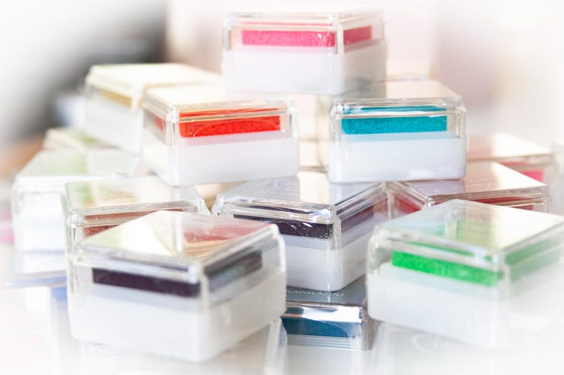 Pode incluir: Uma cole&ccedil;&atilde;o de almofadas de tinta de pl&aacute;stico transparente com almofadas de tinta coloridas dentro. As almofadas de tinta est&atilde;o empilhadas umas sobre as outras e dispostas em forma de pir&acirc;mide.