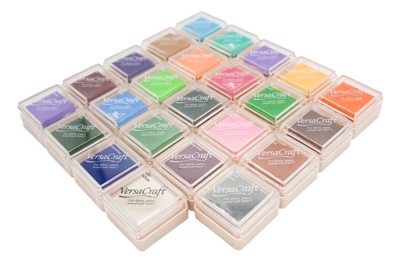 K&ouml;nnte beinhalten: Ein Set mit 24 Stempelkissen in verschiedenen Farben, angeordnet in einem quadratischen Muster. Jedes Kissen ist mit "VersaCraft" beschriftet und in einem durchsichtigen Plastikbeh&auml;lter verpackt.