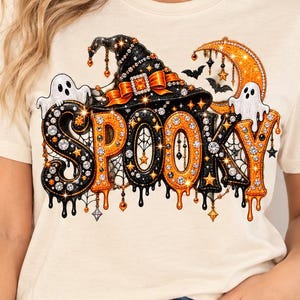 Könnte beinhalten: Ein cremefarbenes Kurzarm-T-Shirt mit einem grafischen Aufdruck des Wortes SPOOKY in großen, stilisierten Buchstaben. Das Design zeigt einen schwarzen Hexenhut, zwei weiße Geister, eine orangefarbene Mondsichel und kleine Fledermäuse mit orangefarbenen Akzenten.