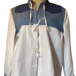 May include: A long-sleeved shirt featuring a white lower section and a dark blue denim upper yoke. The garment includes a stand-up collar, denim-trimmed cuffs, and two white cord ties at the center front for closure. The design is structured and neutral.