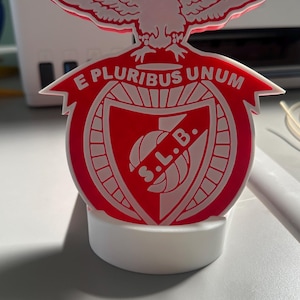 Op de afbeelding: Een rood en wit 3D-geprint embleem met een adelaar bovenop een schild met de tekst E PLURIBUS UNUM en S.L.B. Het embleem is gemonteerd op een ronde witte voet en toont een gedetailleerd grafisch ontwerp met strakke lijnen en heldere kleuren.