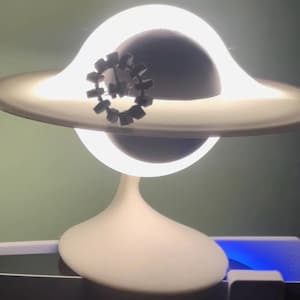 Peut inclure: Une lampe en forme de planète blanche avec un anneau lumineux et un petit modèle de station spatiale circulaire sombre fixé à l'avant. La lampe présente une base incurvée lisse et émet une lumière vive et diffuse depuis l'anneau entourant la sphère.