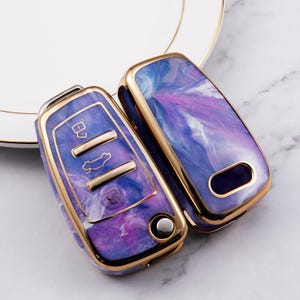 May include: Two car key fob covers featuring a glossy purple, blue, and white marbled pattern with metallic gold-toned edges. The left cover displays three rectangular buttons and a circular button, while the right cover is a solid shell piece.