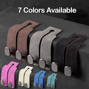 Può includere: Una collezione di sette ganci per poggiatesta auto in vari colori, tra cui marrone, grigio, nero, rosa, beige, blu e turchese. Ogni gancio presenta una cinghia simile alla pelle scamosciata con un attacco in metallo e il testo 7 Colors Available.