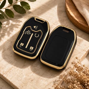 Puede incluir: Dos fundas para llave de coche en negro brillante con borde dorado metálico. Una funda muestra iconos blancos para bloquear, desbloquear, arranque remoto y maletero. La otra es una carcasa negra lisa con bordes dorados y un orificio superior.