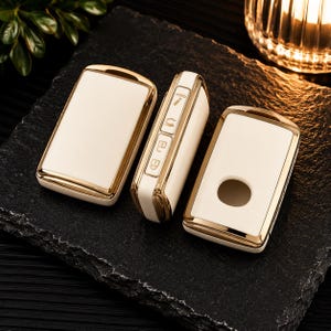 May include: Three white and gold car key fob covers. Two covers are rectangular with rounded corners, one featuring a circular cutout. The third cover is positioned vertically, displaying side buttons with icons for locking, unlocking, and a hold function.