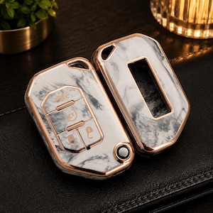 May include: A two-piece car key fob cover featuring a white and grey marble pattern with metallic rose gold trim. The front piece displays moulded button icons for locking, unlocking, and remote start, while the back piece has a rectangular cutout.