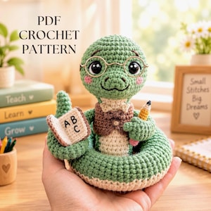 Op de afbeelding: Een groene gehaakte slang met een goudkleurige bril en een bruin vestje. Het figuurtje houdt een klein open boekje met de letters ABC en een geel potlood vast. De tekst PDF CROCHET PATTERN is zichtbaar in de linkerbovenhoek van de afbeelding.