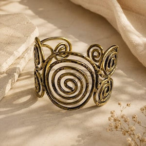 Puede incluir: Un brazalete ancho de metal en tono latón antiguo, que presenta una serie de motivos en espiral interconectados. La superficie del metal tiene un acabado martillado y texturizado, creando un patrón repetitivo de bobinas circulares en toda la banda.