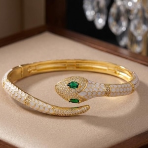 Könnte beinhalten: Ein goldfarbener Schlangen-Armreif mit weißen Emaille-Schuppen, einem mit kleinen klaren Kristallen besetzten Kopf und zwei marquise-geschliffenen grünen Steinen als Augen. Das klappbare Design bildet eine durchgehende Schlangenform.