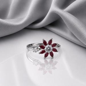 Peut inclure: Une bague argentée ornée d'une pierre centrale ronde transparente entourée de huit pierres rouge foncé taille marquise disposées en fleur, avec une petite pierre transparente supplémentaire fixée sur l'anneau près du motif floral.