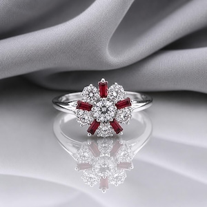 Peut inclure: Une bague argentée ornée d'une pierre centrale ronde transparente, entourée d'un motif floral composé de pierres rondes transparentes alternées avec des pierres rouges rectangulaires de taille baguette sur un anneau poli.