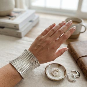 Può includere: Un ampio bracciale a fascia in argento con una texture a coste verticali. Il bracciale presenta una finitura metallica lucida e una forma curva che avvolge il polso. Il design è composto da linee in rilievo che si ripetono sulla superficie.