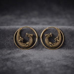 May include: A pair of circular hoop earrings crafted from antique brass-toned metal. Each earring features a detailed, textured dragon figure coiled within the center of the hoop, showcasing intricate scale patterns and a defined head and tail.