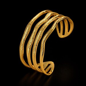 Puede incluir: Un brazalete tipo brazalete de tono dorado con cuatro bandas paralelas y onduladas. La superficie de las bandas tiene un acabado texturizado y granulado, mientras que los extremos del brazalete son lisos y redondeados. El diseño es abierto.