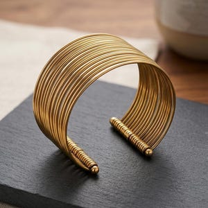 Può includere: Un bracciale a fascia color oro composto da molteplici fili metallici sottili e paralleli che si curvano per formare una forma circolare ampia e aperta. Le estremità dei fili sono fissate da una barra cilindrica scanalata con una punta sferica.