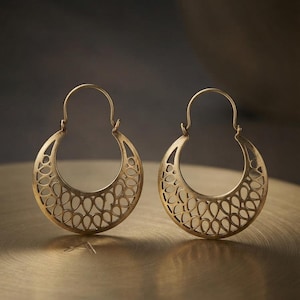 Peut inclure: Une paire de créoles dorées présentant un design en forme de croissant avec un motif ajouré complexe composé de découpes répétitives en forme de goutte d'eau. Les boucles d'oreilles ont une attache en fil courbé et une finition métallique mate.