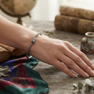 May include: A silver-toned metal bracelet featuring a textured, scale-like pattern along the band and a detailed snake head design at the center. The bracelet is a flexible, open-ended cuff style that wraps around the wrist with a polished finish.