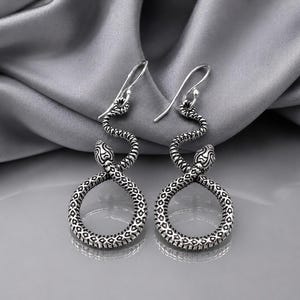 May include: A pair of silver-toned drop earrings featuring a snake design. Each earring depicts a serpent with a textured, scaled pattern, coiled into a circular shape with the head positioned at the top, suspended from a simple silver hook.