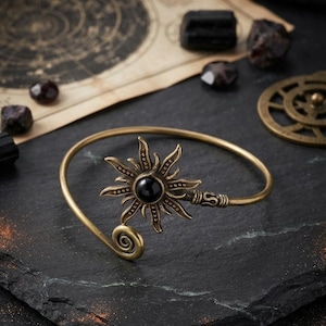May include: A brass-toned metal cuff bracelet featuring a central sunburst motif with a smooth, round black stone at its core. The bracelet has a thin, open-ended band that terminates in a spiral design on one side and a textured, coiled detail.