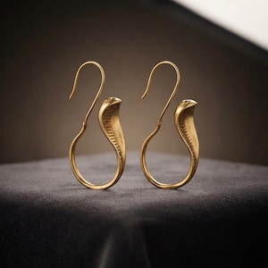 May include: A pair of gold-toned earrings shaped like stylised cobras. Each earring features a circular hoop base that transitions into an upright, flared cobra head with textured detailing along the neck, finished with a curved hook for attachment.