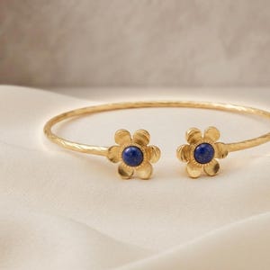 May include: A gold-toned open cuff bracelet featuring a textured band and two flower-shaped terminals. Each flower has six petals and a central circular dark blue stone inlay. The bracelet has a gap between the two floral ends.