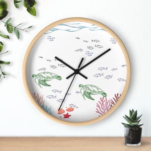 Könnte beinhalten: Eine runde Wanduhr mit hellem Holzrahmen zeigt ein weißes Zifferblatt mit grünen Meeresschildkröten, kleinen blauen Fischen, roter Koralle und einem Seestern. Drei schwarze Zeiger sind mittig auf dem Zifferblatt mit Wellenmuster angebracht.