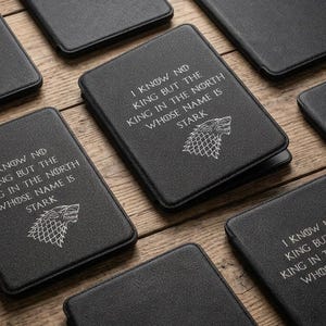 May include: Black rectangular e-reader cases with a textured finish. Each case features the white printed text I KNOW NO KING BUT THE KING IN THE NORTH WHOSE NAME IS STARK and a graphic of a wolf head emblem centered below the text.
