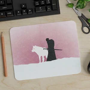 May include: A rectangular mouse pad featuring a graphic illustration of a dark silhouette of a person standing with a sword next to a white wolf. The background is a textured dusty rose pink color with a white snowy landscape at the bottom edge.