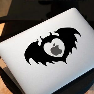 Op de afbeelding: Een zilverkleurige laptop met een zwarte vinylsticker op de deksel. De sticker toont een hartvorm met vleermuisachtige vleugels aan de zijkanten. In het hart zit een uitsparing die het grijze Apple-logo van de laptop eronder onthult.