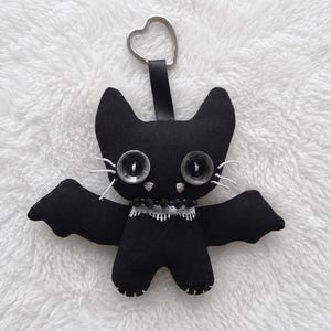 May include: A black fabric plush bat keychain featuring two large dark button eyes, white stitched whiskers, a beaded collar with a white lace trim, and a silver-toned heart-shaped metal ring attached to the top by a black fabric strap.