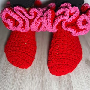 May include: A pair of bright red crocheted slippers featuring a thick, ruffled cuff in a contrasting light pink yarn. The slippers are constructed with a dense, textured stitch pattern and are worn over black leggings, covering the feet completely.