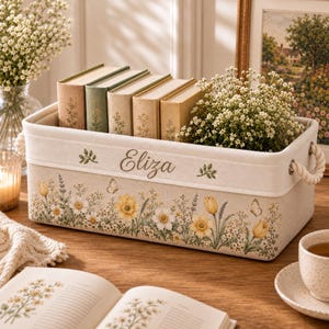 Könnte beinhalten: Ein rechteckiger Aufbewahrungskorb aus beigem Stoff mit einem Blumenmuster aus gelben und weißen Blüten, grünen Blättern und Schmetterlingen. Der Name Eliza ist in Schreibschrift auf der Vorderseite eingestickt. Der Korb hat weiße Seilgriffe.