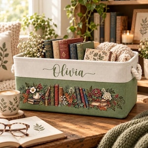 Op de afbeelding: Een rechthoekige stoffen opbergmand in saliegroen en crème, voorzien van de naam Olivia in cursief schrift. De buitenkant toont een illustratie van gestapelde boeken, herfstbladeren en bloemstukken. De mand heeft witte touwgrepen.