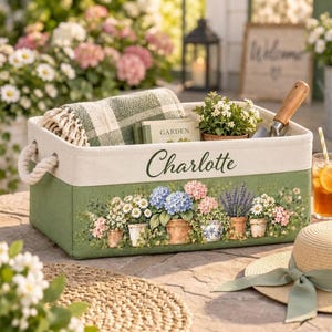 May include: A rectangular fabric storage bin with rope handles, featuring the name Charlotte printed in dark green script. The base is sage green with a colourful floral print of potted flowers, and a green plaid blanket is tucked inside.
