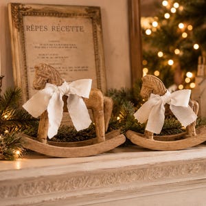 May include: Two light brown wooden rocking horse figurines, each adorned with a white fabric bow tied around the neck. The horses feature carved details on the mane and saddle area, standing on curved rockers, displayed as decorative ornaments.