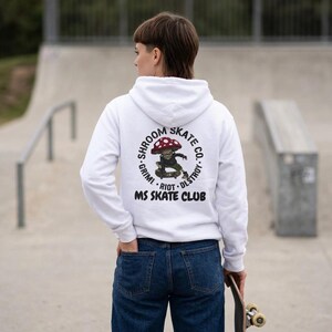 May include: A white hooded sweatshirt featuring a graphic on the back of a mushroom character skateboarding. The circular text reads SHROOM SKATE CO. GRIM! RIOT! DESTROY! and MS SKATE CLUB is printed below the graphic in bold black lettering.