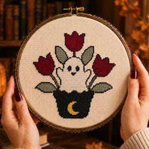 May include: A cross-stitch embroidery piece in a circular wooden hoop featuring a white ghost sitting inside a black pot decorated with a yellow crescent moon. The ghost is surrounded by three dark red tulips with sage green leaves on cream fabric.