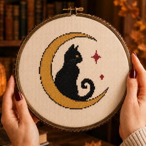 Peut inclure: Un tambour à broder circulaire contenant un motif au point de croix sur un tissu crème. Le motif représente un chat noir assis sur un croissant de lune jaune, accompagné de deux petites étoiles rouge foncé à quatre branches sur le côté.