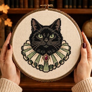 May include: A cross-stitch embroidery piece in a circular wooden hoop featuring a black cat with green eyes wearing a pleated, light green and white ruffled collar with a small, central red gemstone accent, stitched on a cream-colored fabric.