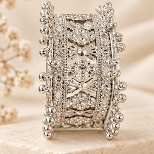 May include: A wide silver-toned cuff bracelet featuring an intricate openwork design with repeating floral and geometric patterns. The edges are lined with rows of small, dangling silver spheres that create a textured, beaded appearance.