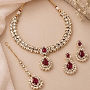 May include: A gold-toned jewelry set consisting of a double-strand necklace, a pair of teardrop earrings, and a maang tikka. Each piece features clear white stones and a central deep red teardrop-shaped stone surrounded by small white accents.