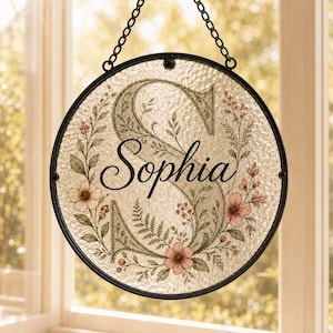 May include: A circular glass suncatcher featuring a large, muted green letter S as a background. The name Sophia is printed in black script across the center. The design is decorated with delicate illustrations of pink flowers and green foliage.