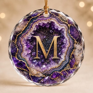 May include: A circular glass ornament featuring a printed purple and white amethyst geode pattern with gold-colored veining. A large, gold-colored serif letter M is centered on the design. The ornament has a faceted edge and a gold hanging cord.