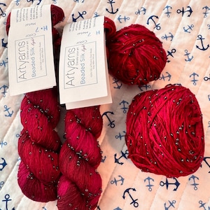 Puede incluir: Dos madejas y un ovillo de hilo Artyarns Beaded Silk Light de color rojo intenso, cada uno con pequeñas cuentas oscuras integradas en la fibra de seda. Cada madeja incluye una etiqueta de papel blanco con el texto Artyarns Beaded Silk light.